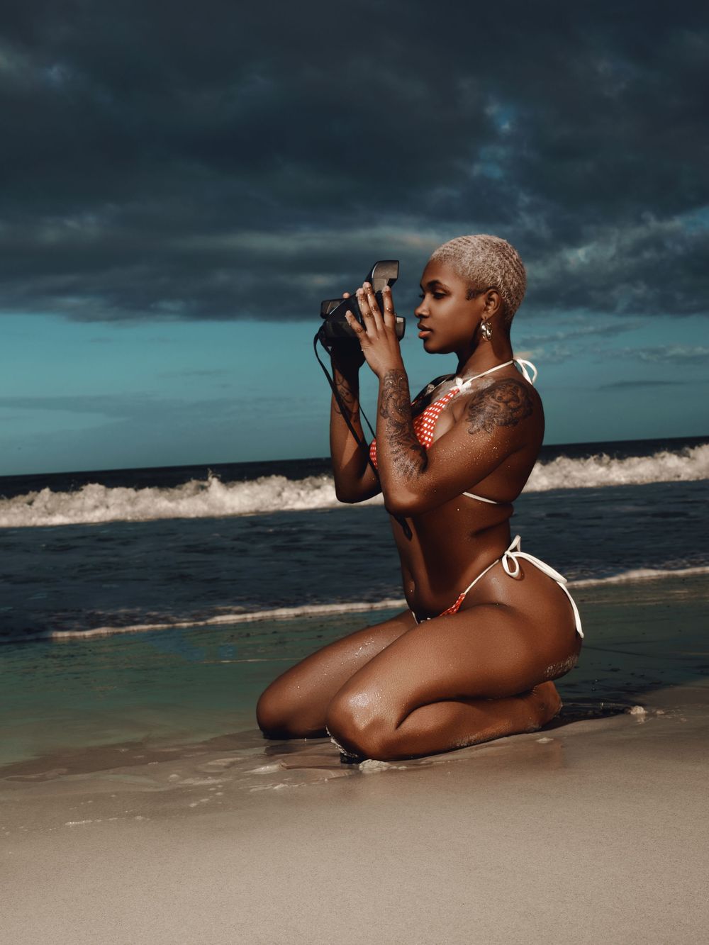 A figure in a white swimsuit poses artistically on a beach at sunset with dramatic stormy clouds overhead.