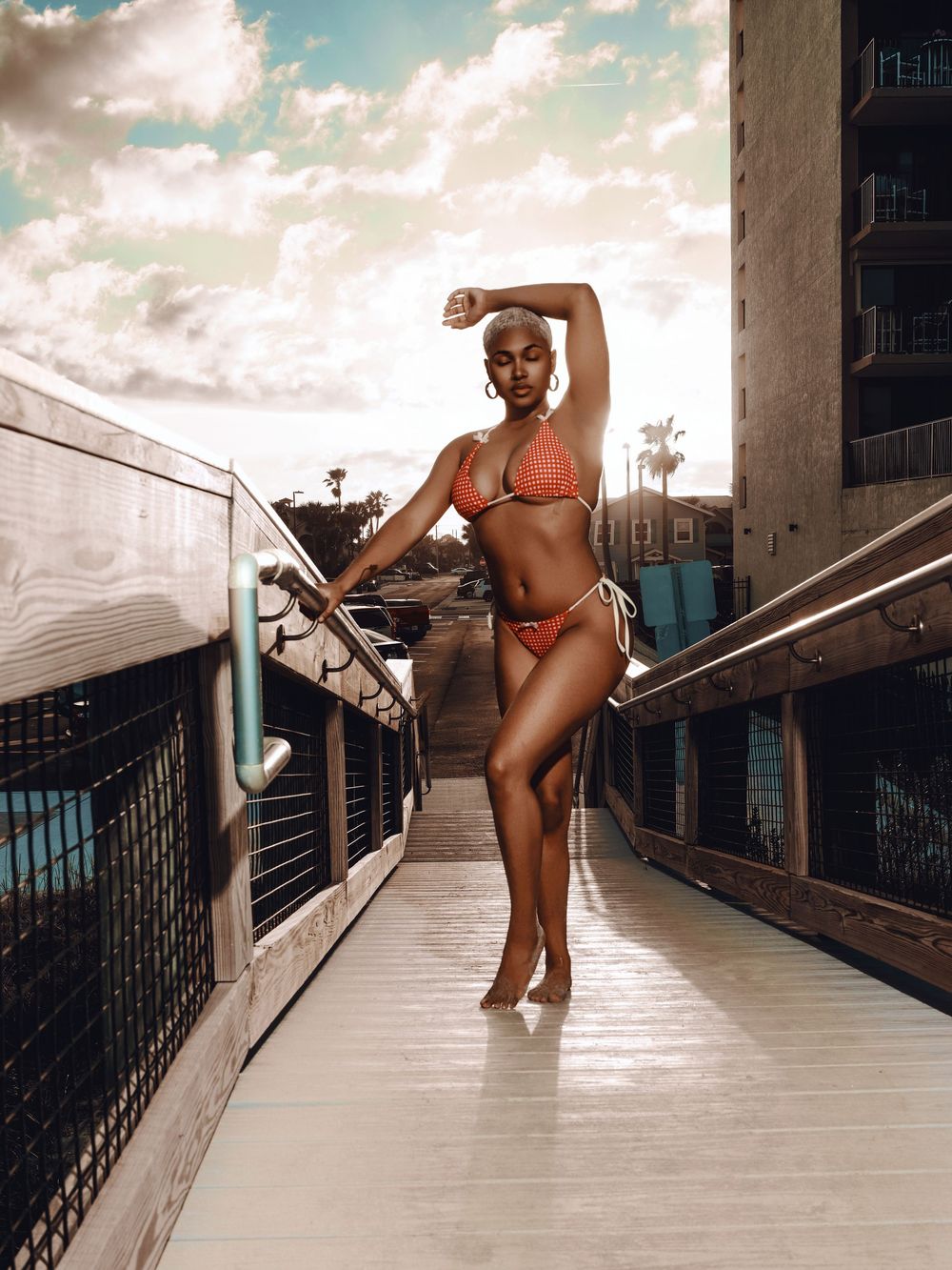 Athletic figure in a red bikini strikes a graceful pose on an elevated walkway against a cloudy sky and urban backdrop.