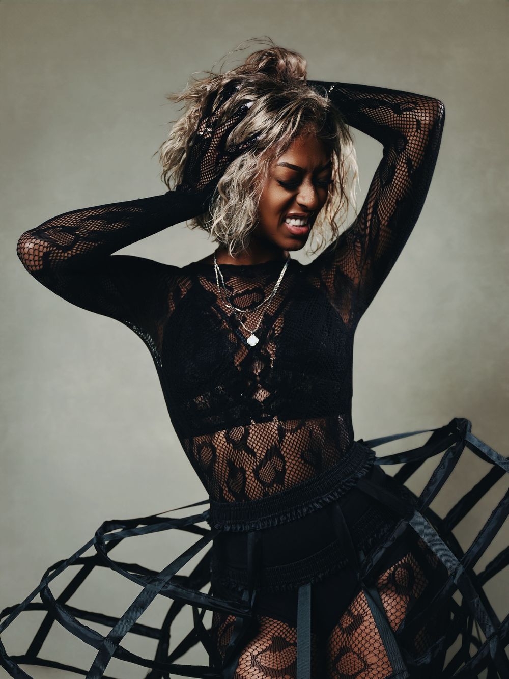 Woman in black lace bodysuit and cage skirt poses dramatically with hands in blonde curly hair.