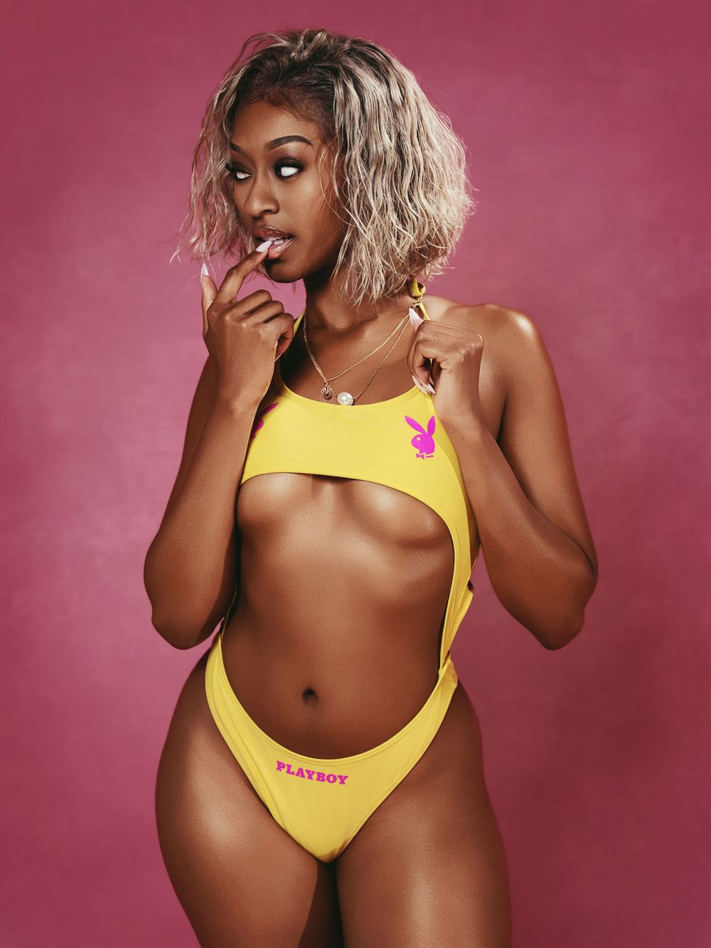 Woman in yellow Playboy cutout swimsuit posing against pink background.