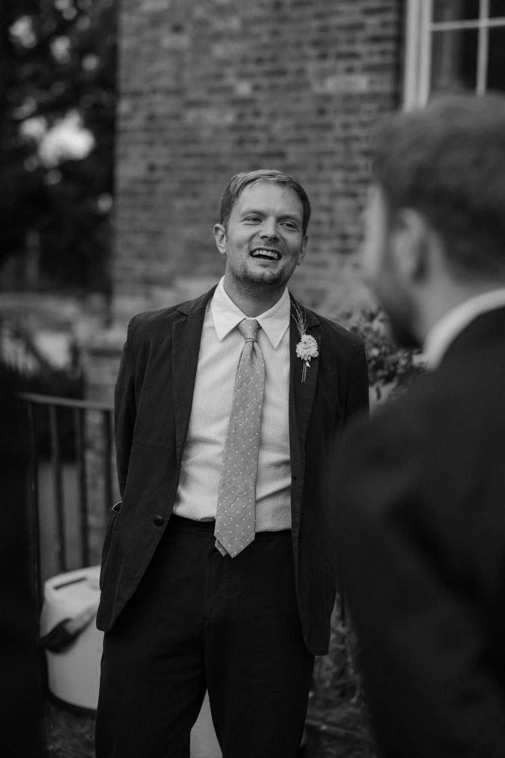 A person in a dark suit and tie shares a candid laughing moment at an outdoor event in black and white photography.