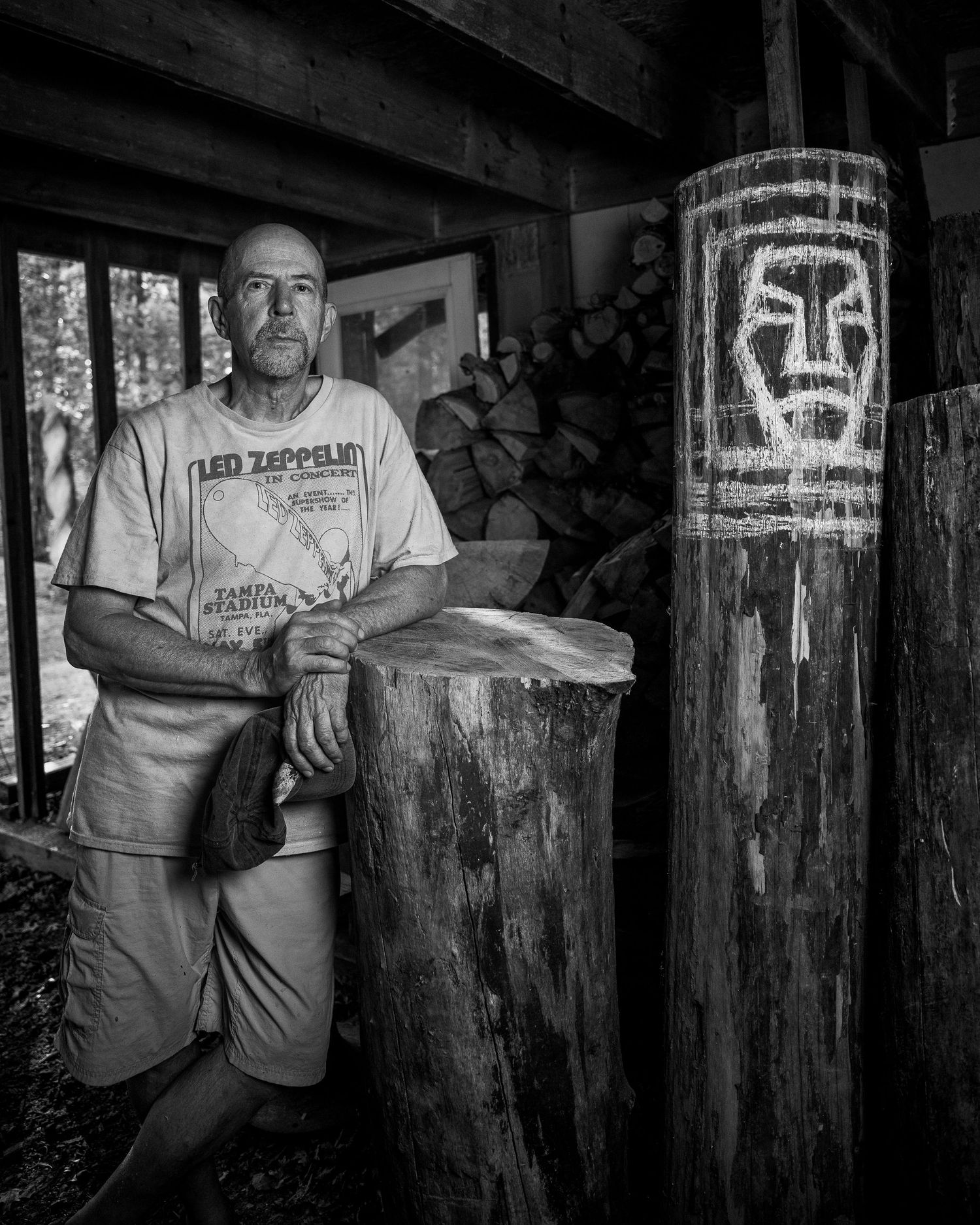 Black and white portrait of a person in a Led Zeppelin shirt standing next to a wooden post with tribal art carving.