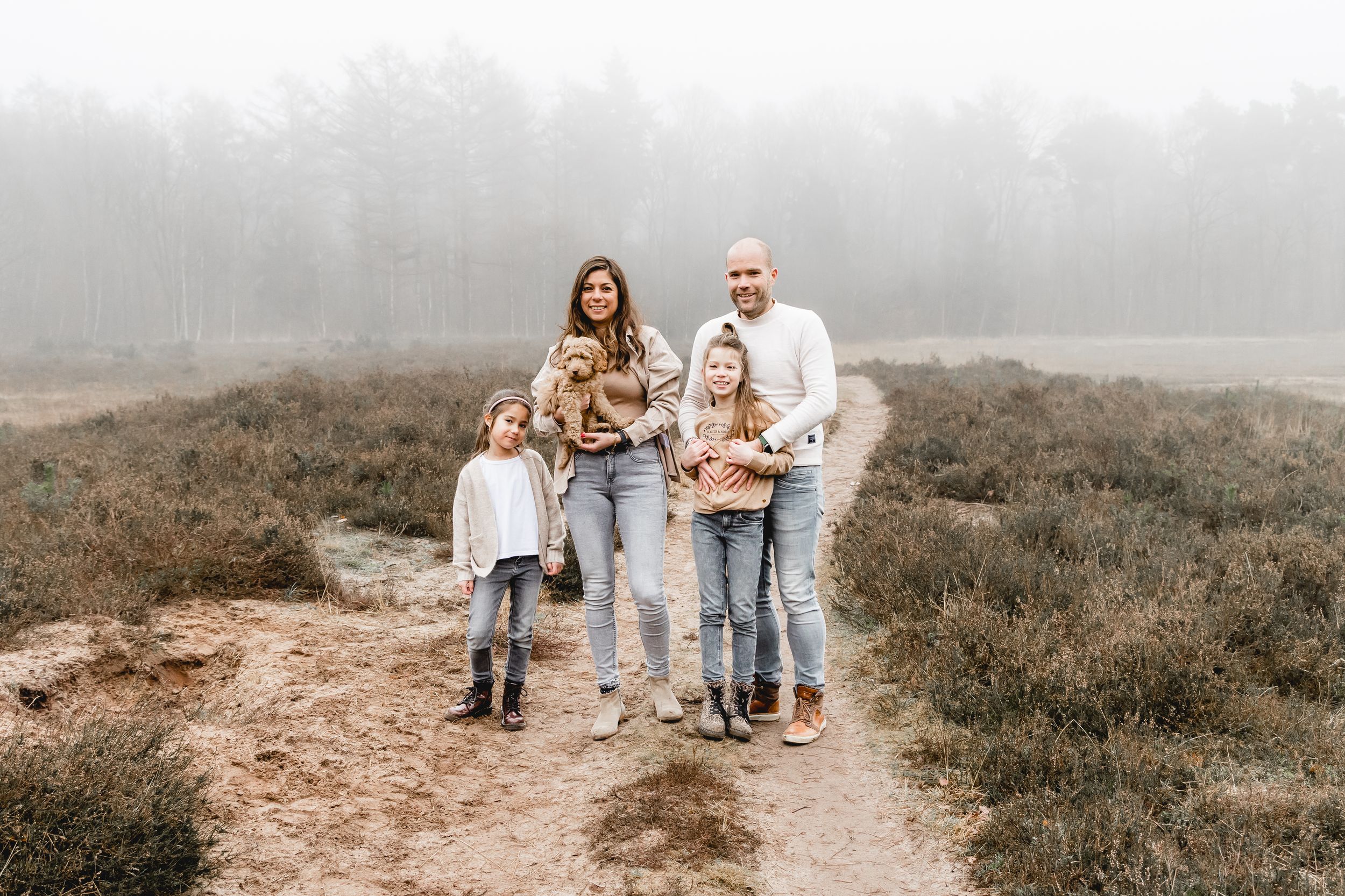 A family dressed in neutral colors poses together on a misty beach with sand dunes in the background.