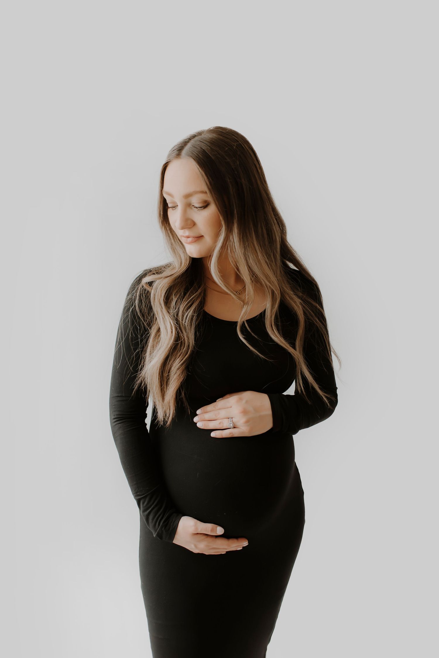 Woman in black dress poses against white background while holding her pregnant belly.
