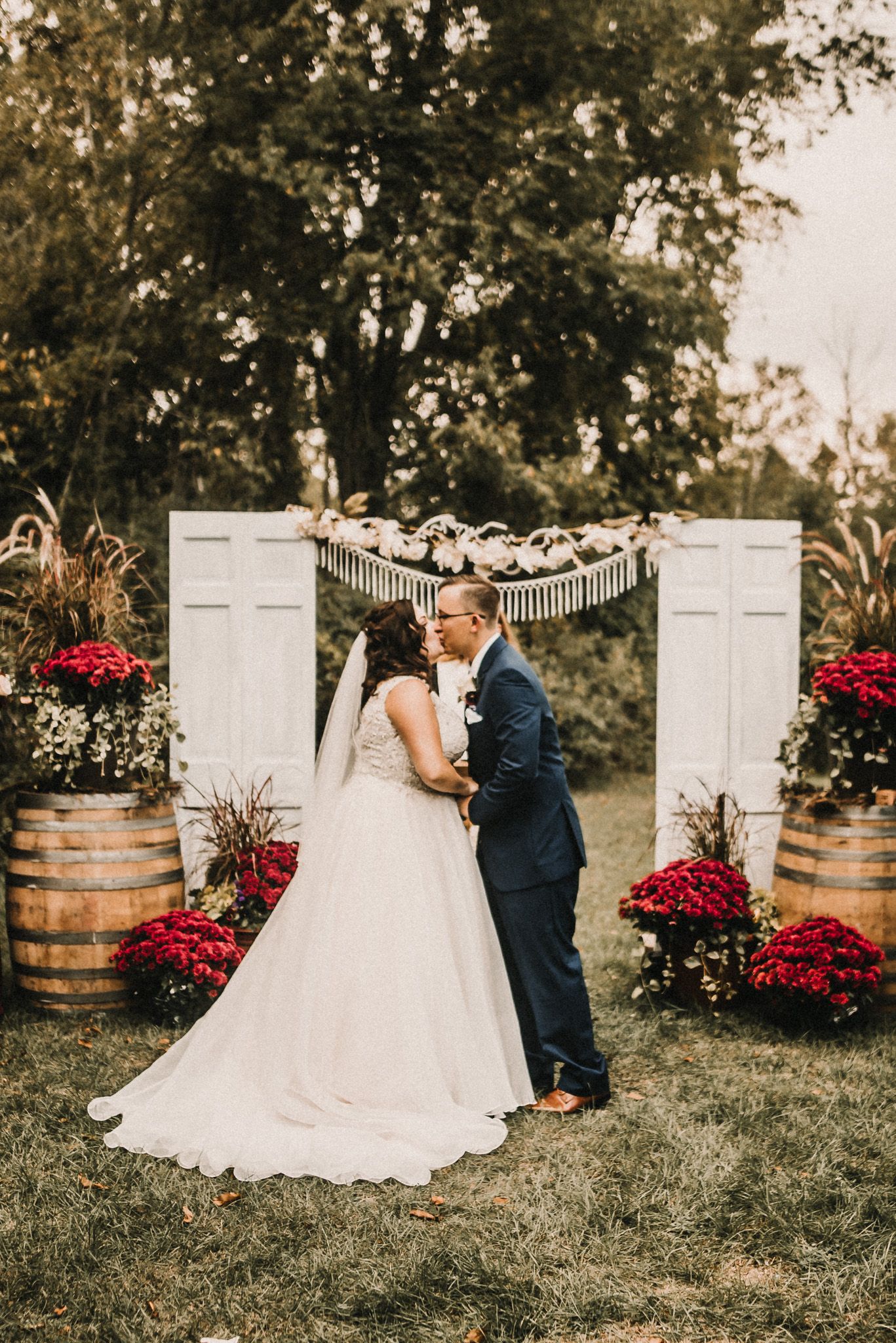 A romantic outdoor wedding scene with decorative white doors, wine barrels, and red flowers in a rustic garden setting.