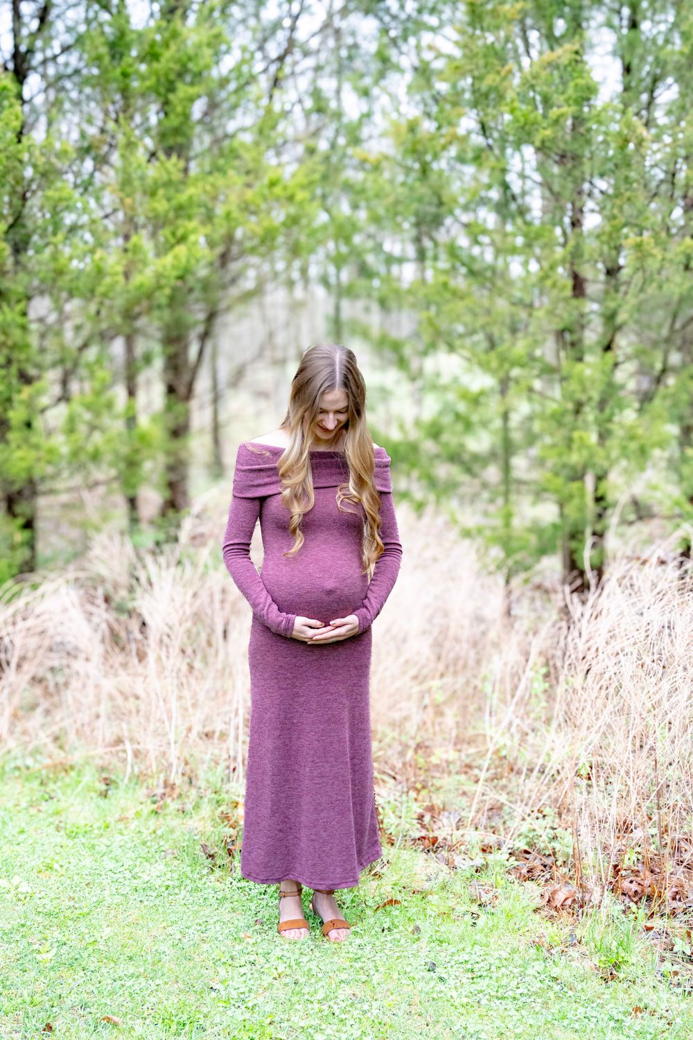 A person in a purple sweater dress stands in a grassy field surrounded by tall dried grass and trees.