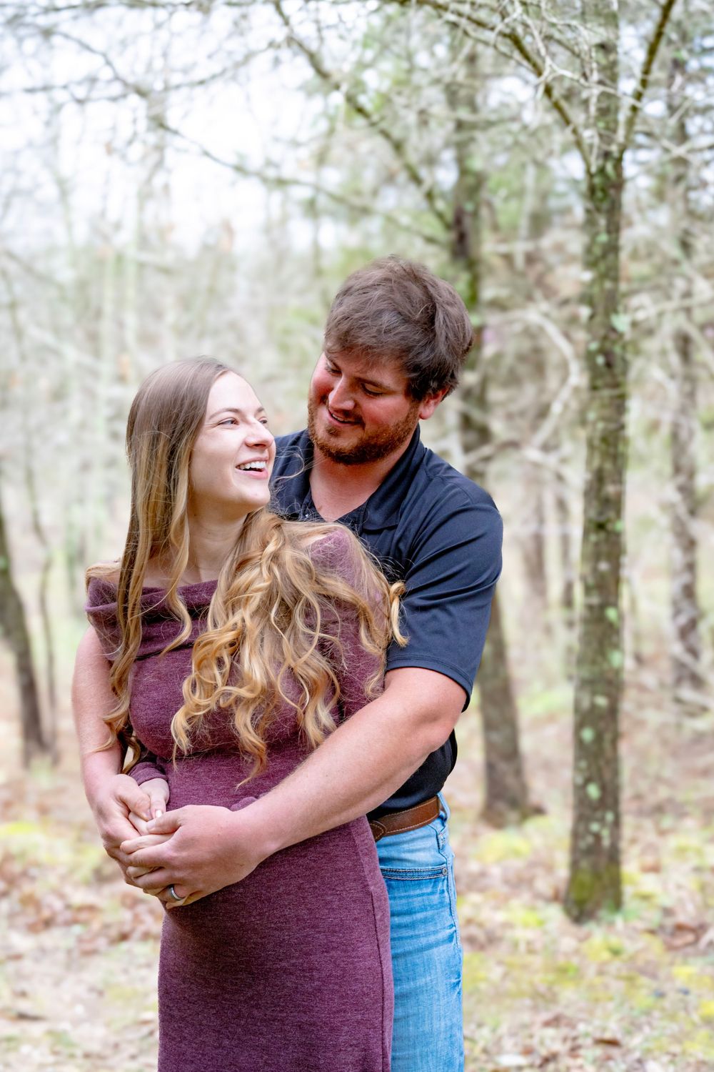 A couple shares a joyful embrace while standing in a wooded forest setting on a bright day.