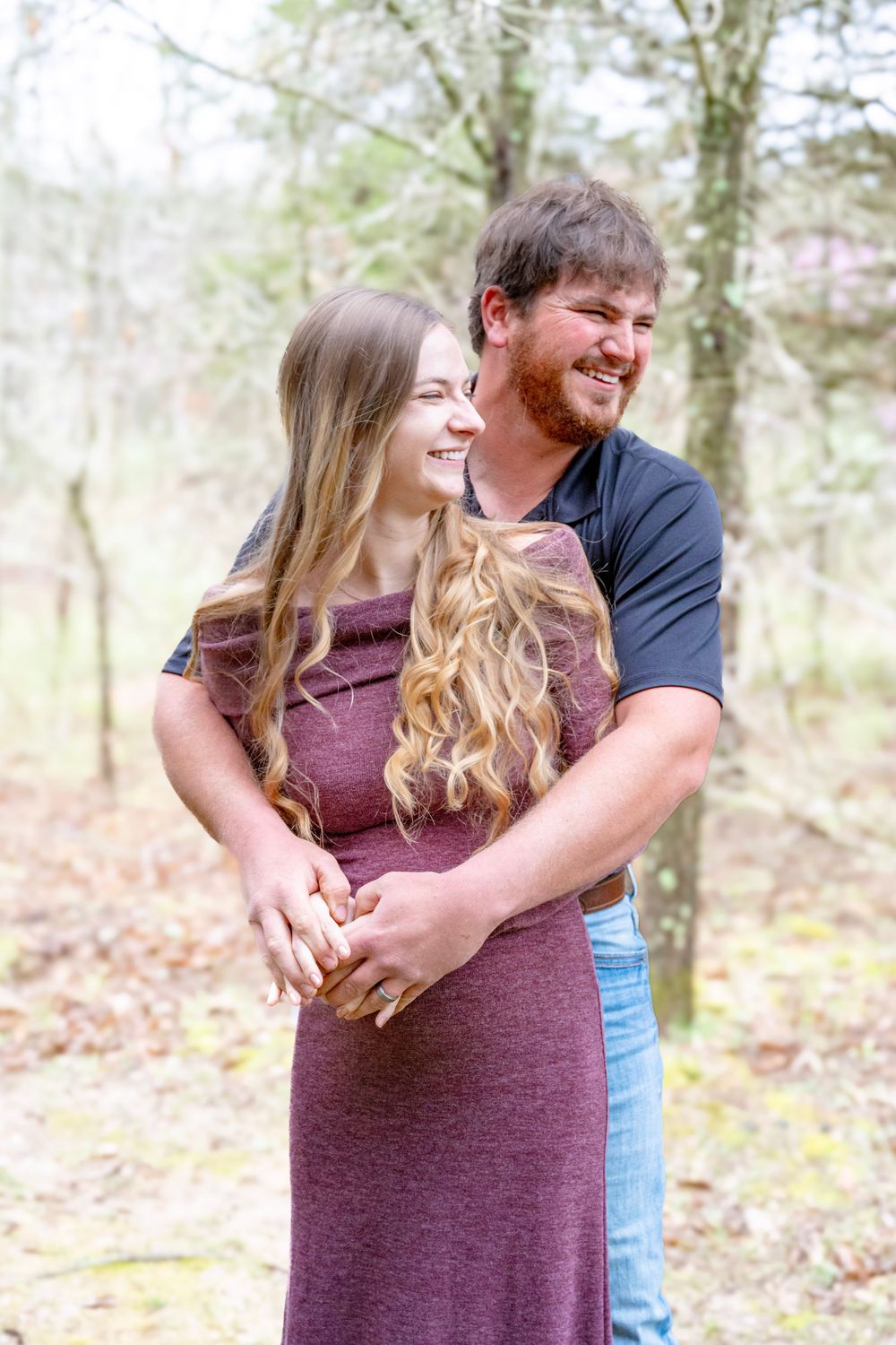 A couple embraces outdoors in a wooded setting, with the woman wearing a burgundy dress and the man in a navy shirt.
