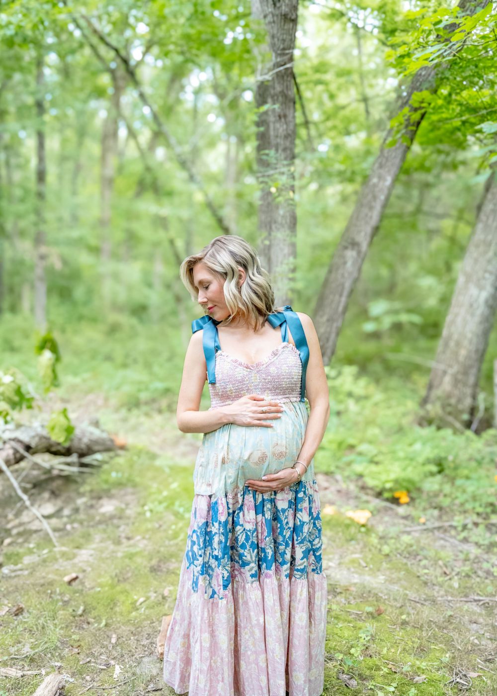 A person in a floral dress stands in a lush green forest setting during a summer day.