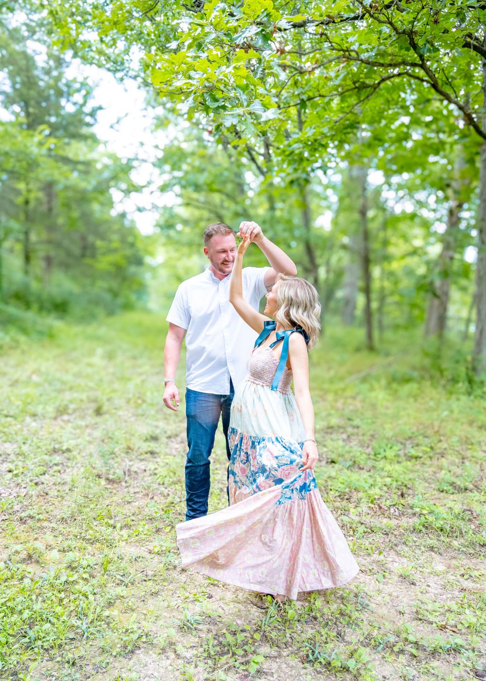 A couple in white and floral outfits dance together on a grassy hillside surrounded by lush green trees.