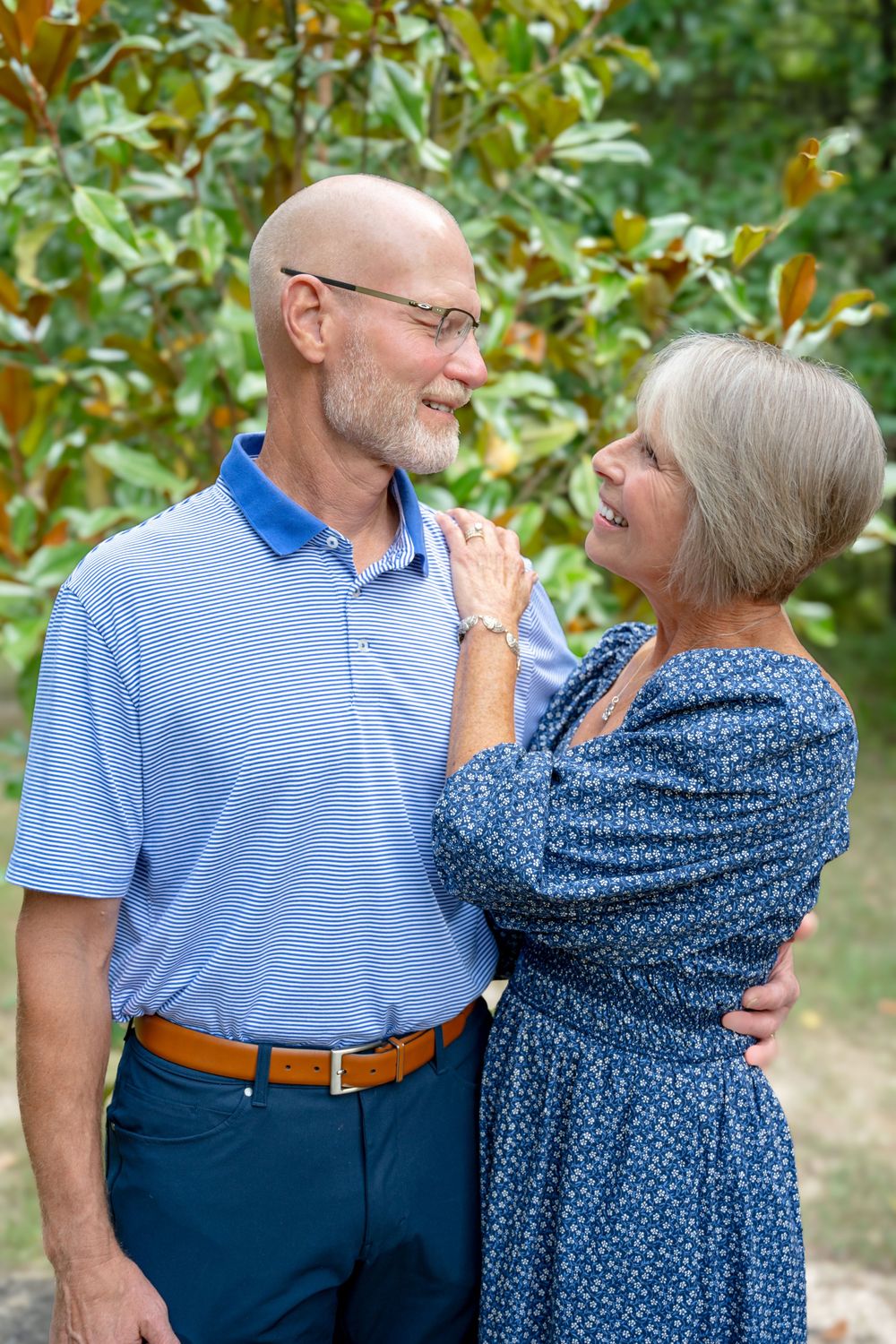 A loving senior couple embraces and smiles at each other while standing outdoors among green foliage.