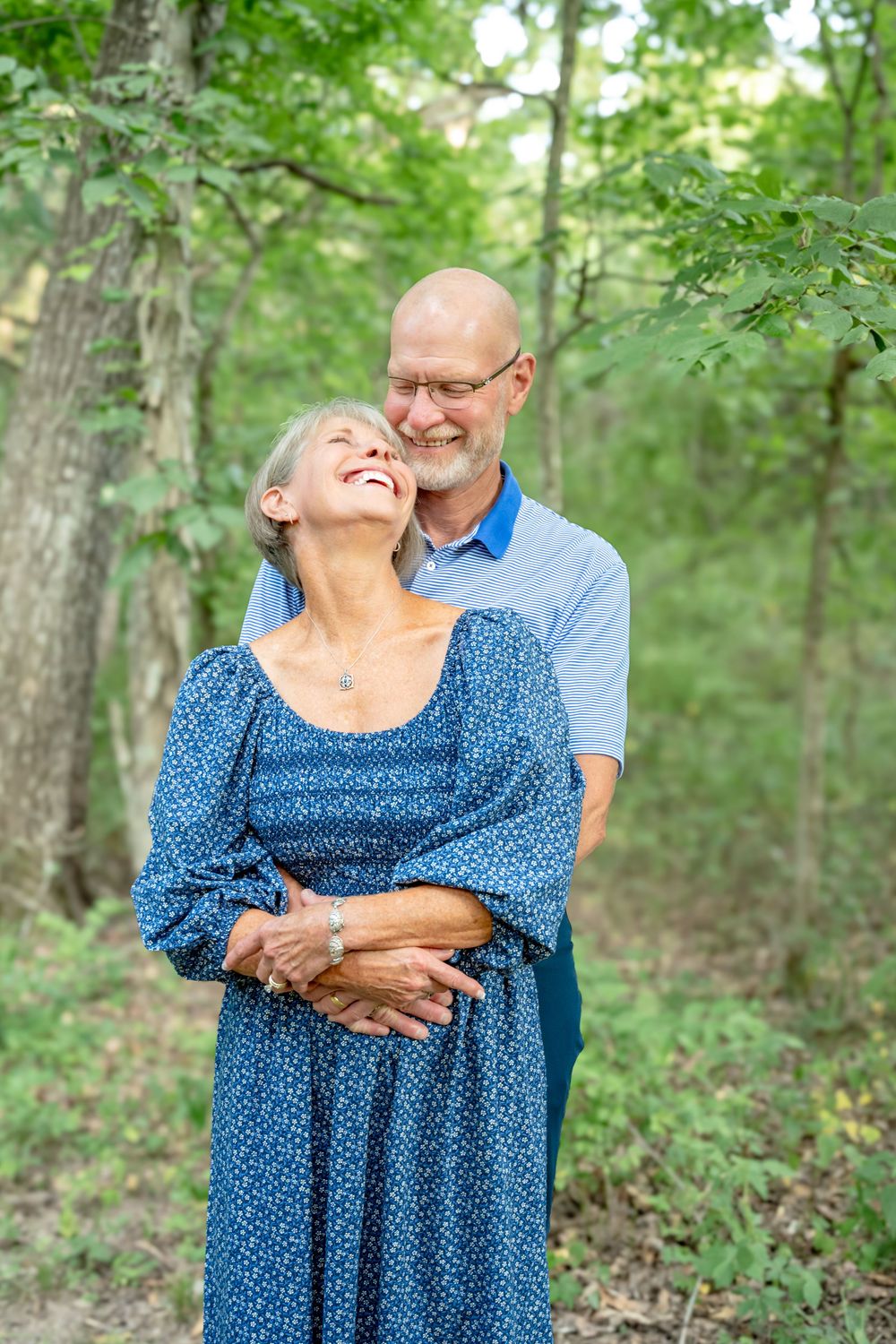 A couple embracing and laughing together in a sunlit forest setting while wearing coordinating blue outfits.