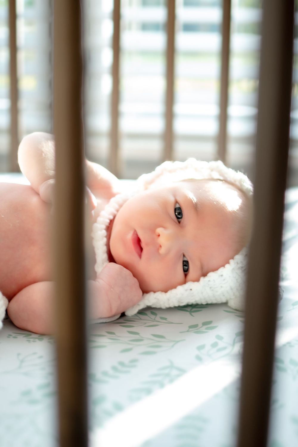 Baby lying in crib with white bedding seen through wooden slats in soft natural lighting.