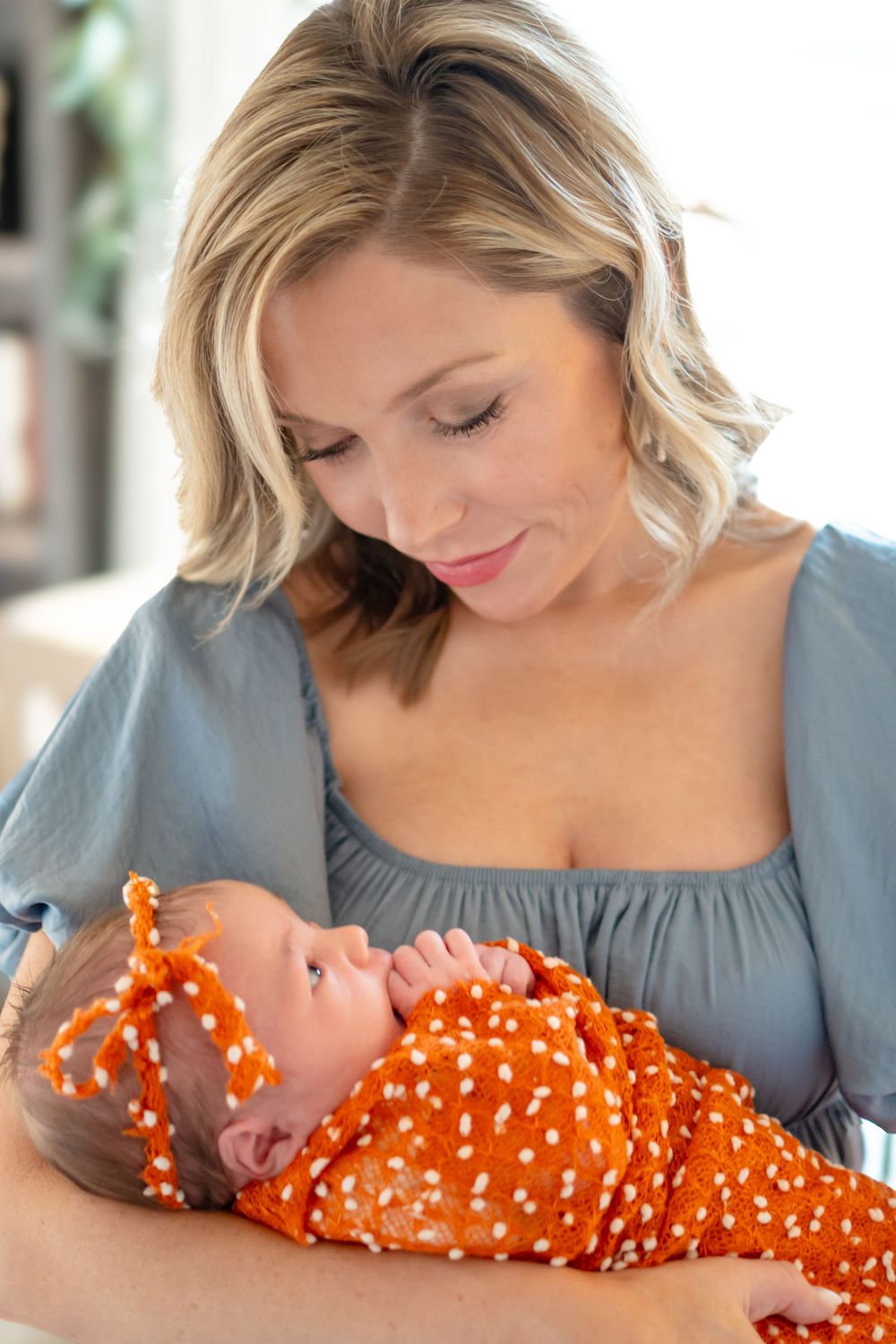 Mother in gray dress lovingly holds newborn baby wearing orange polka dot outfit.