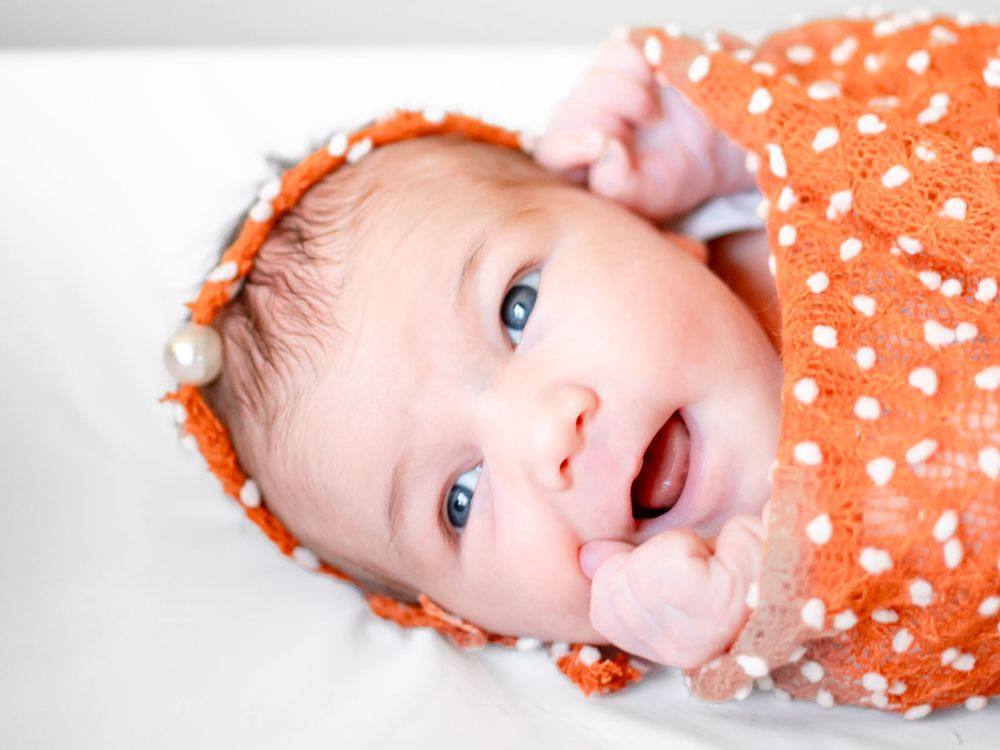 Baby wearing orange floral outfit smiles and plays while lying on side against matching orange background.