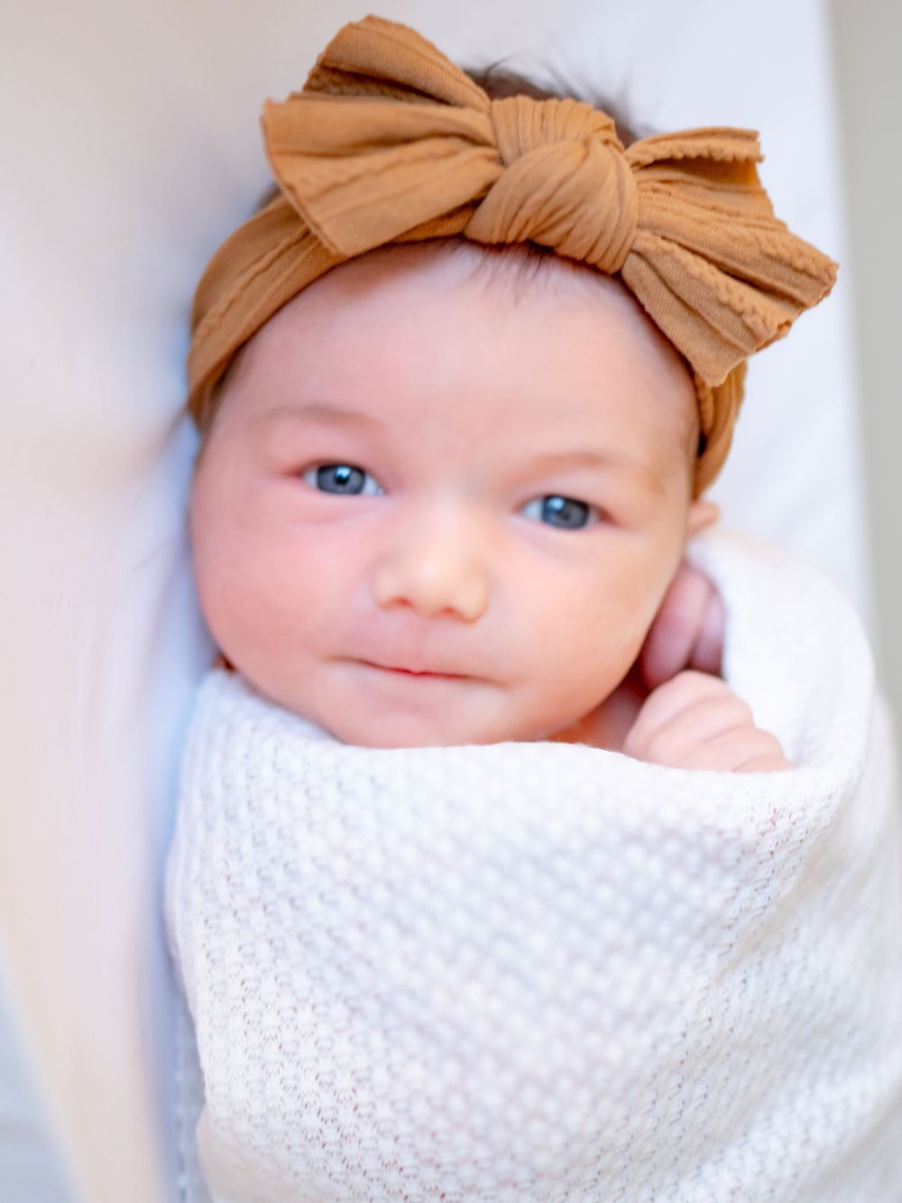 A baby wearing a tan bow headband is swaddled in a white knit blanket.