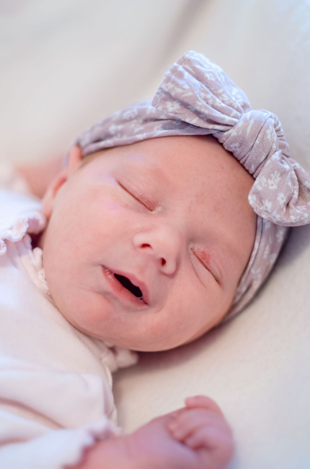Sleeping newborn baby wearing a soft gray bow headband rests peacefully on white bedding.