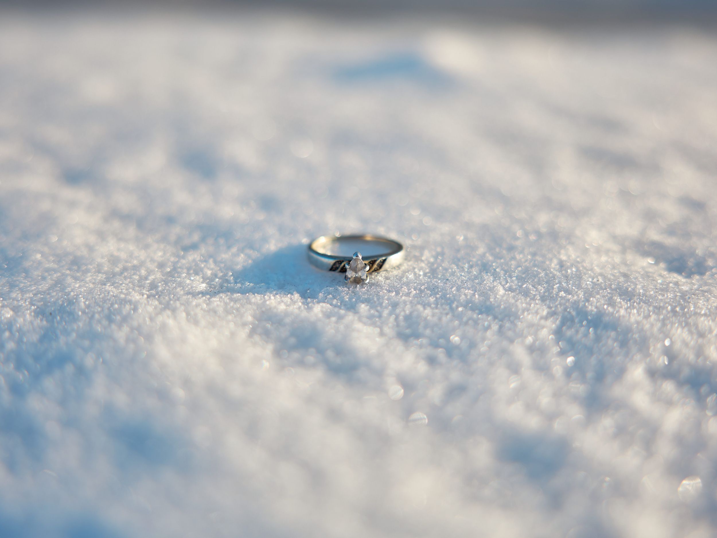 A diamond engagement ring sparkles in pristine white snow with soft bokeh effect and blue winter lighting.