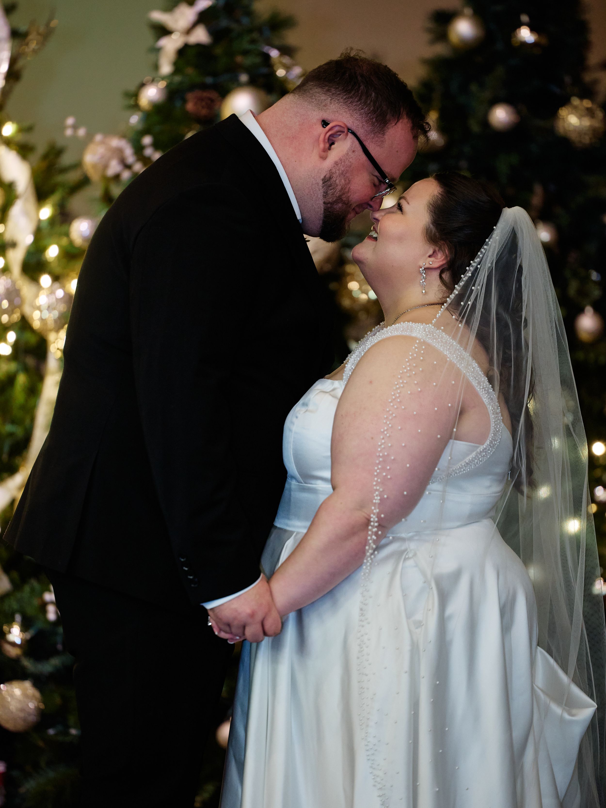 A romantic wedding moment captured in front of a Christmas tree with twinkling lights and ornaments.