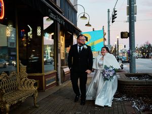 A wedding couple walks down a city street at dusk with string lights illuminating the urban scene.