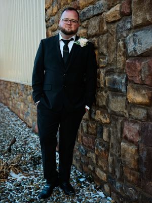 A person in a black suit stands against a rustic stone wall with gravel ground during a winter day.