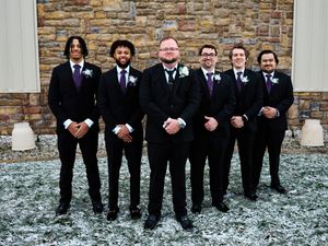 Groomsmen in matching black suits and ties stand together in a line against a stone wall on a snowy winter day.