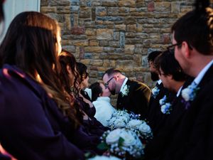Wedding party gathers in a line against stone wall holding white bouquets at dusk.