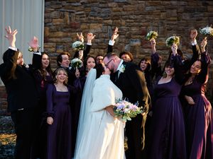 A wedding party in purple dresses celebrates joyfully outside a stone church wall with raised hands.