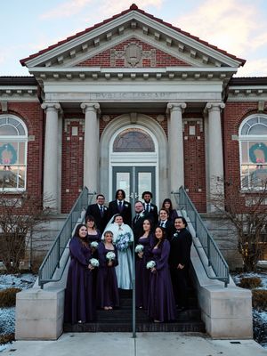 A wedding party poses on the steps of a red brick building with white columns and arched windows in winter.