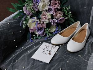 A wedding bouquet with purple roses and white ballet flats next to a 'Mr & Mrs' card on gray fabric.