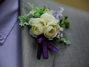 A delicate boutonniere with cream roses and purple accents tied with a sparkly purple ribbon against a gray background.
