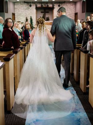A flowing white wedding dress with a long train trails behind as people sit in church pews during a ceremony.