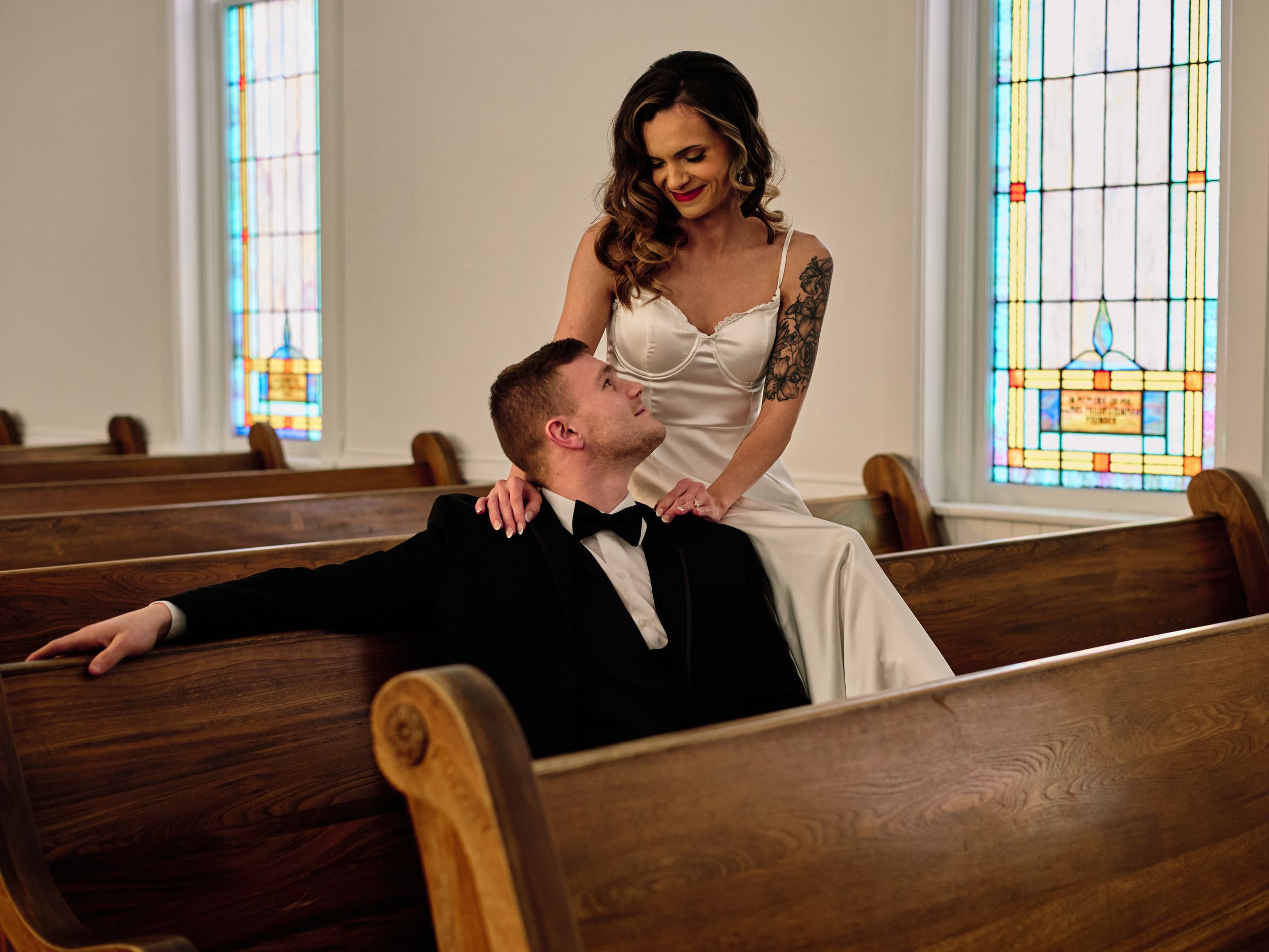 A bride and groom share an intimate moment together in a church pew with stained glass windows in the background.