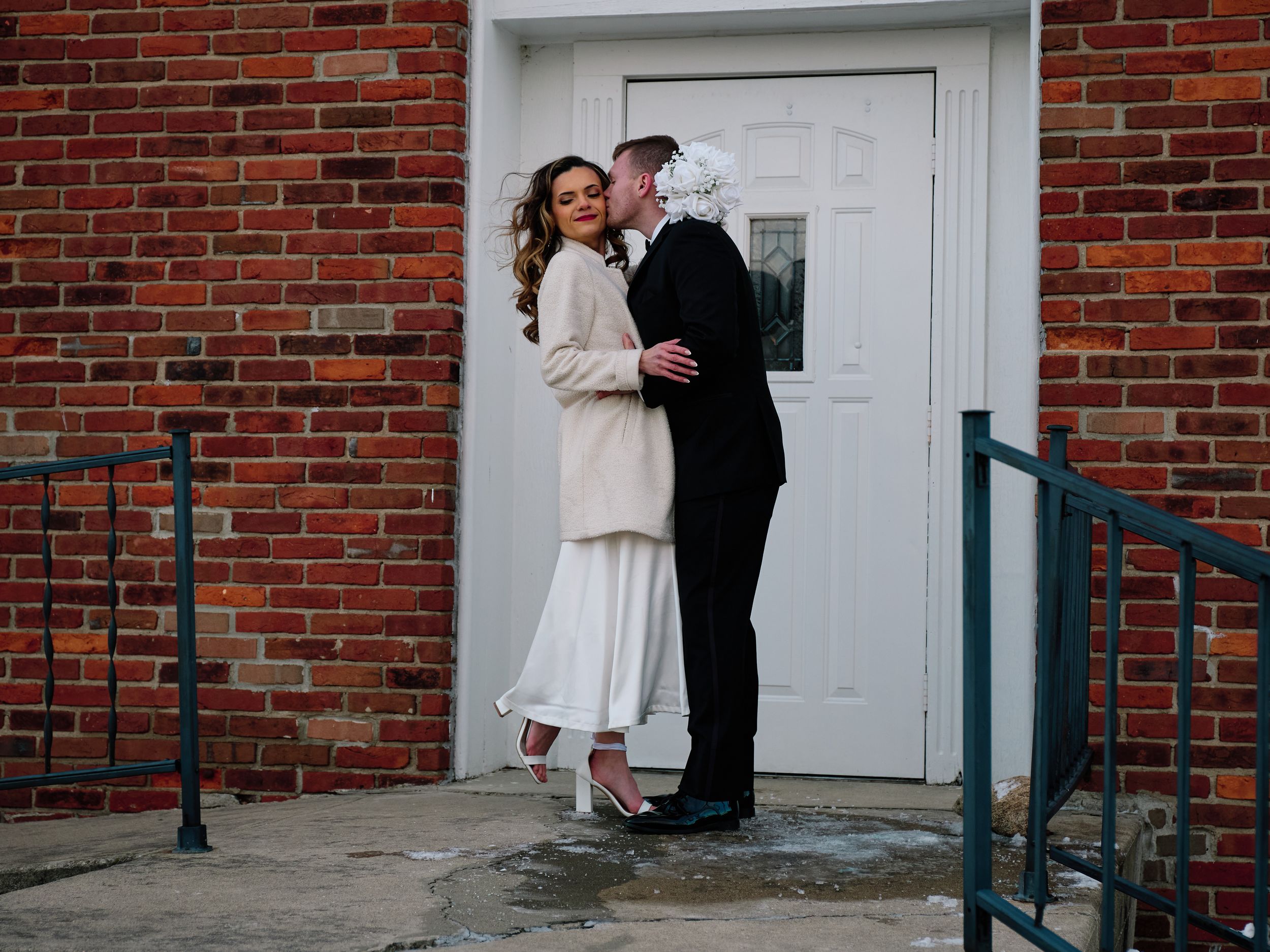 A couple shares a romantic moment in front of a white door on a red brick building entrance.