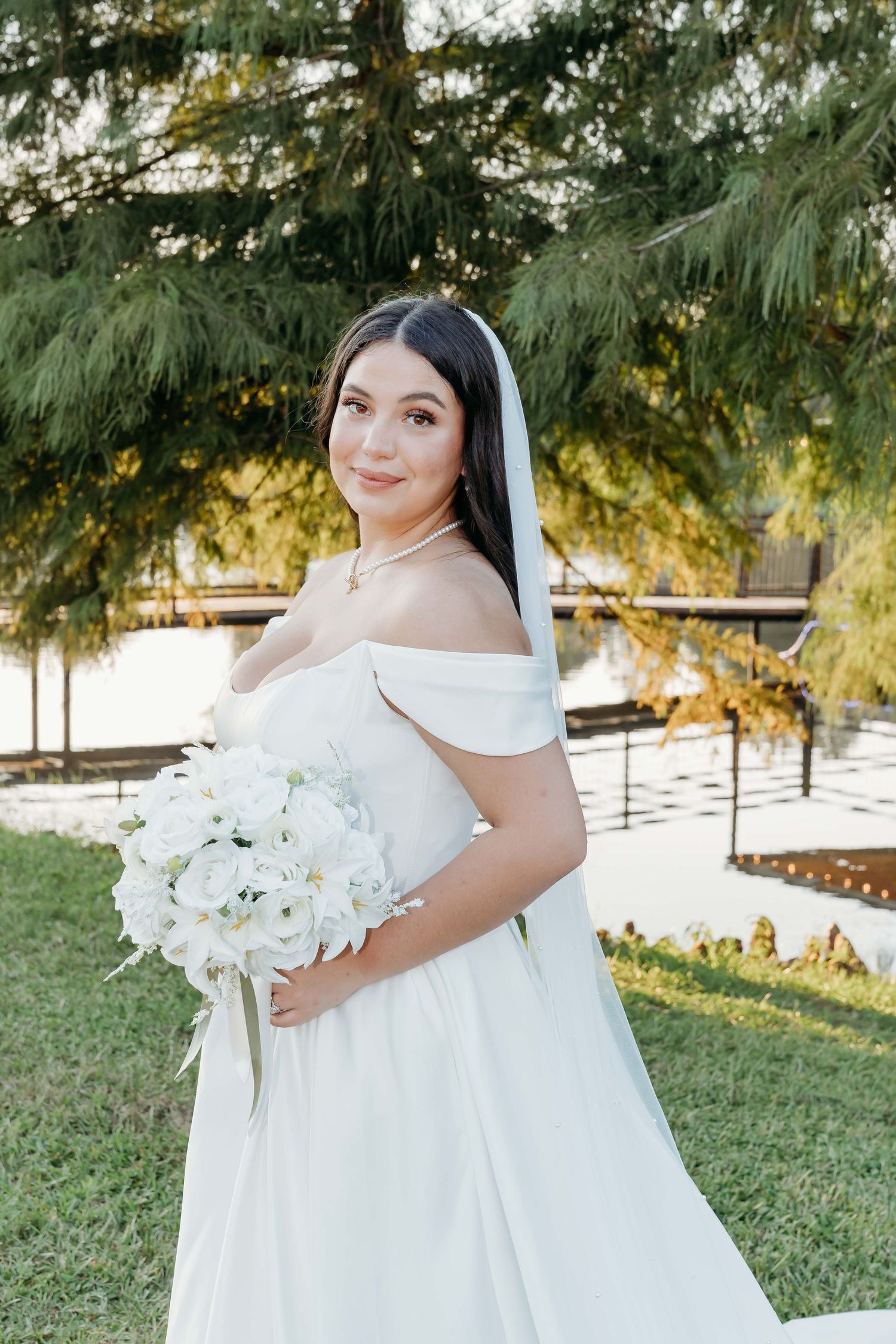 A bride in a flowing white wedding dress poses outdoors with a bouquet of white orchids against a natural backdrop.