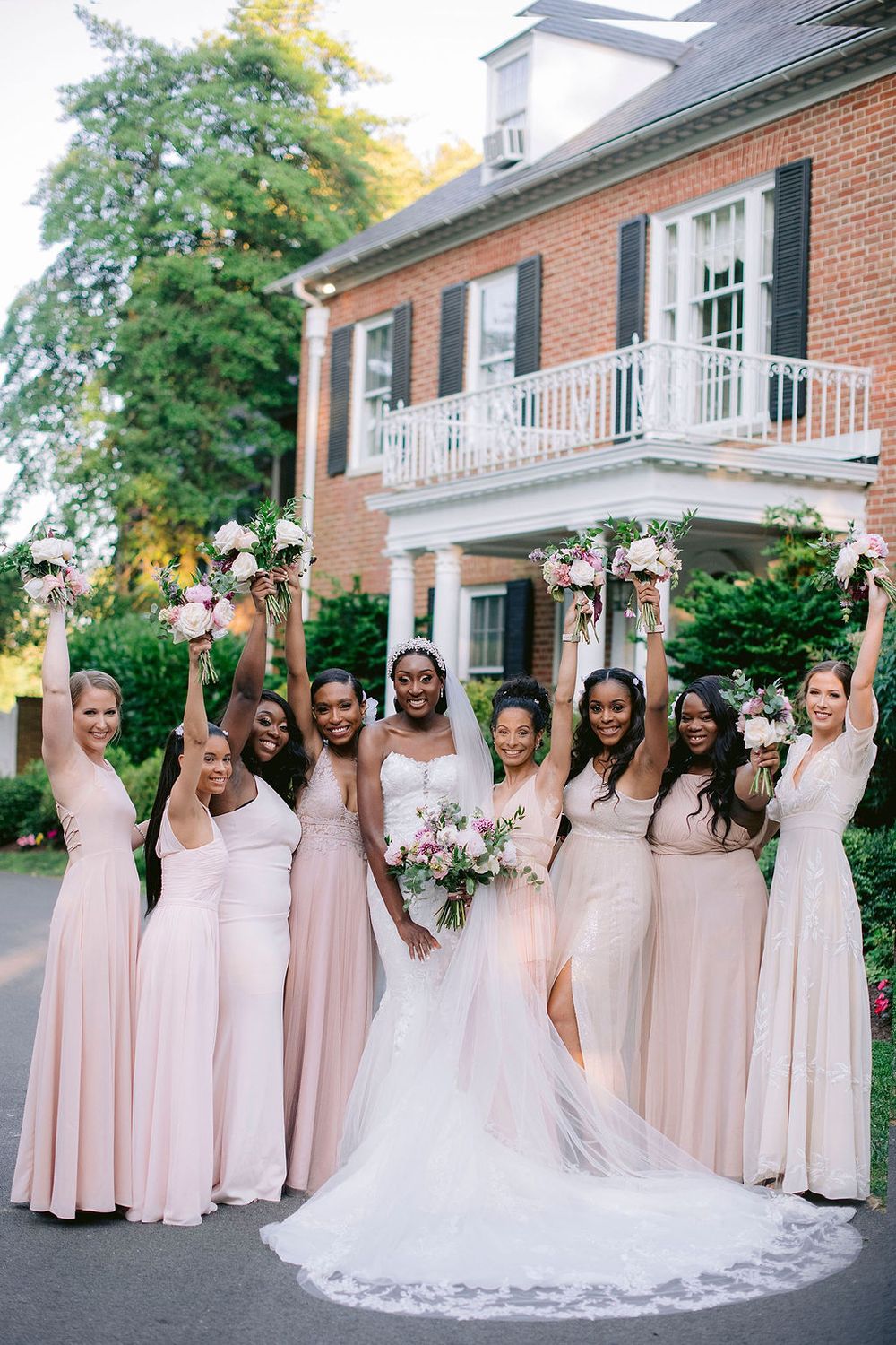 A wedding party wearing blush pink bridesmaid dresses poses together outside a brick building on a sunny day.