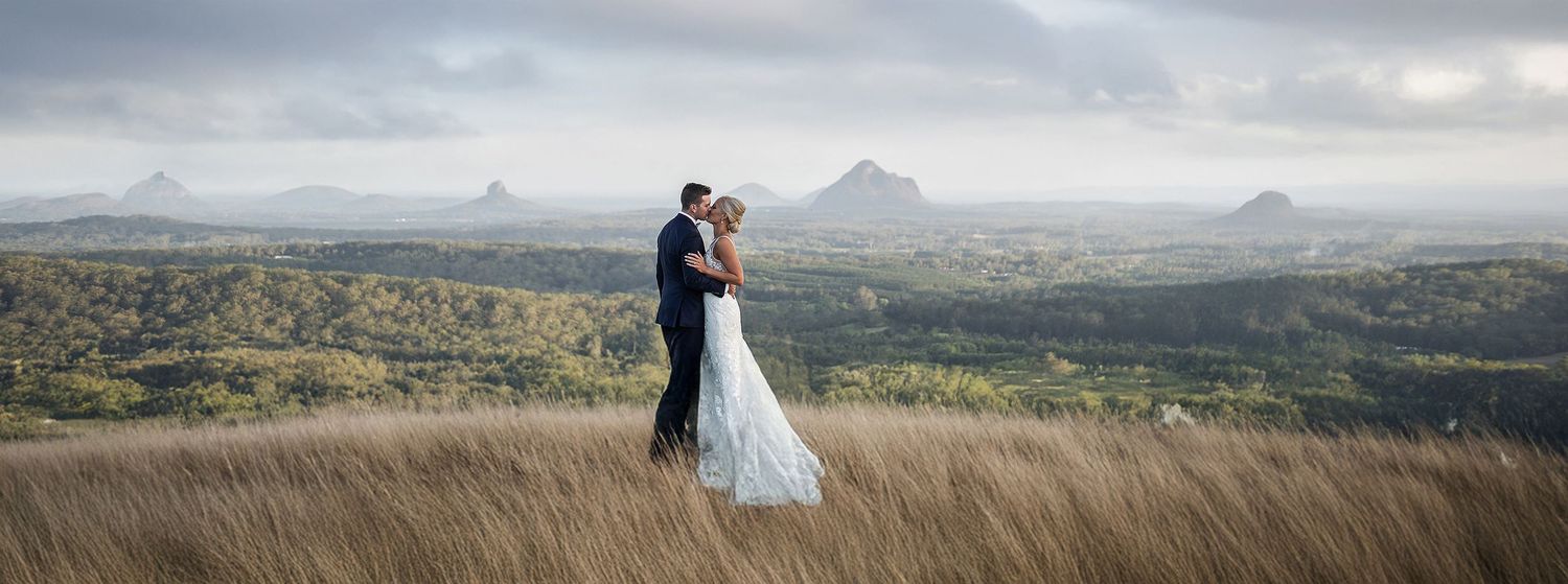 Bride and groom embracing on a hilltop with stunning Glasshouse Mountains panoramic backdrop.