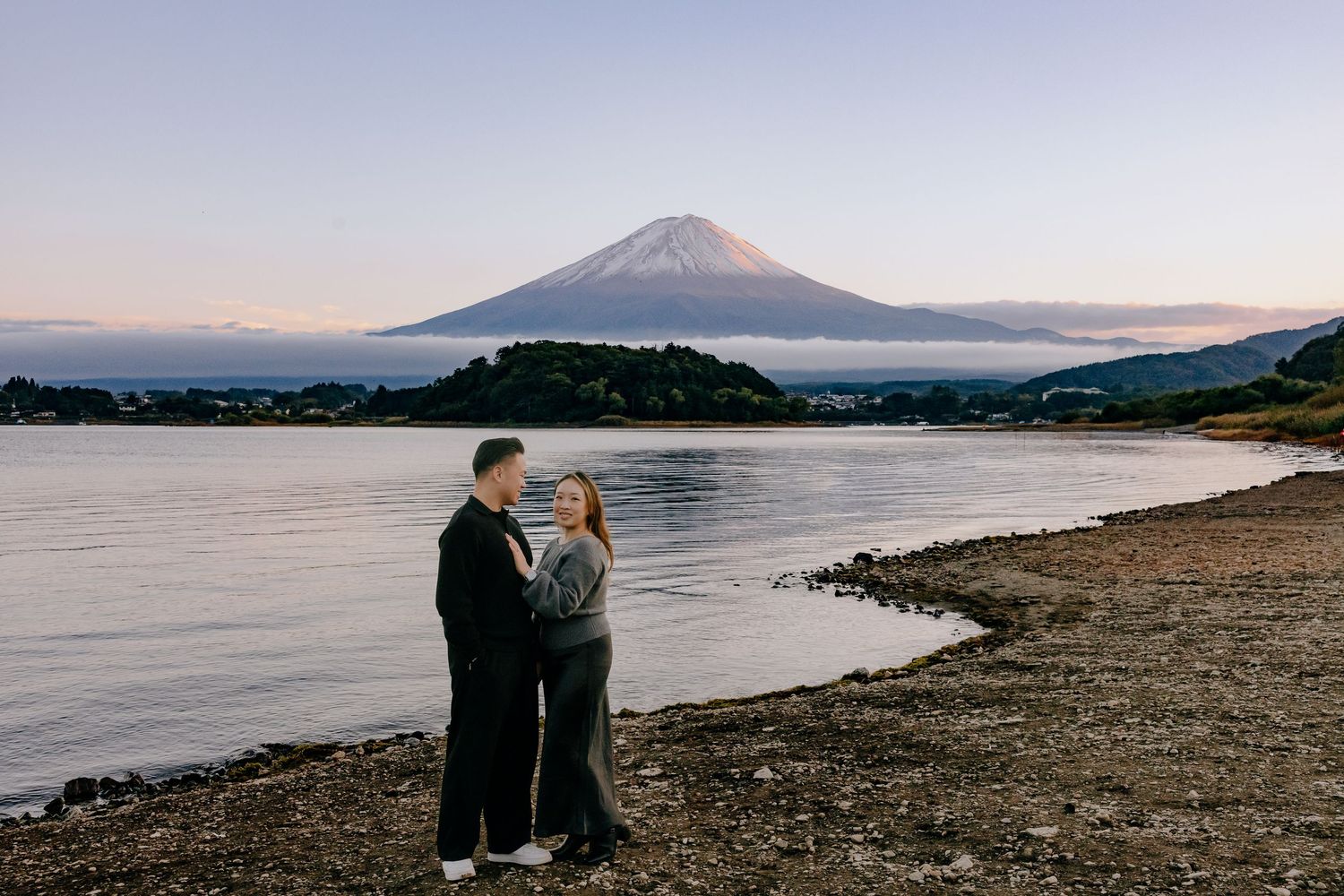 Mount Fuji rises majestically over a tranquil lake shore at sunset with its snow-capped peak reflecting in the water.
