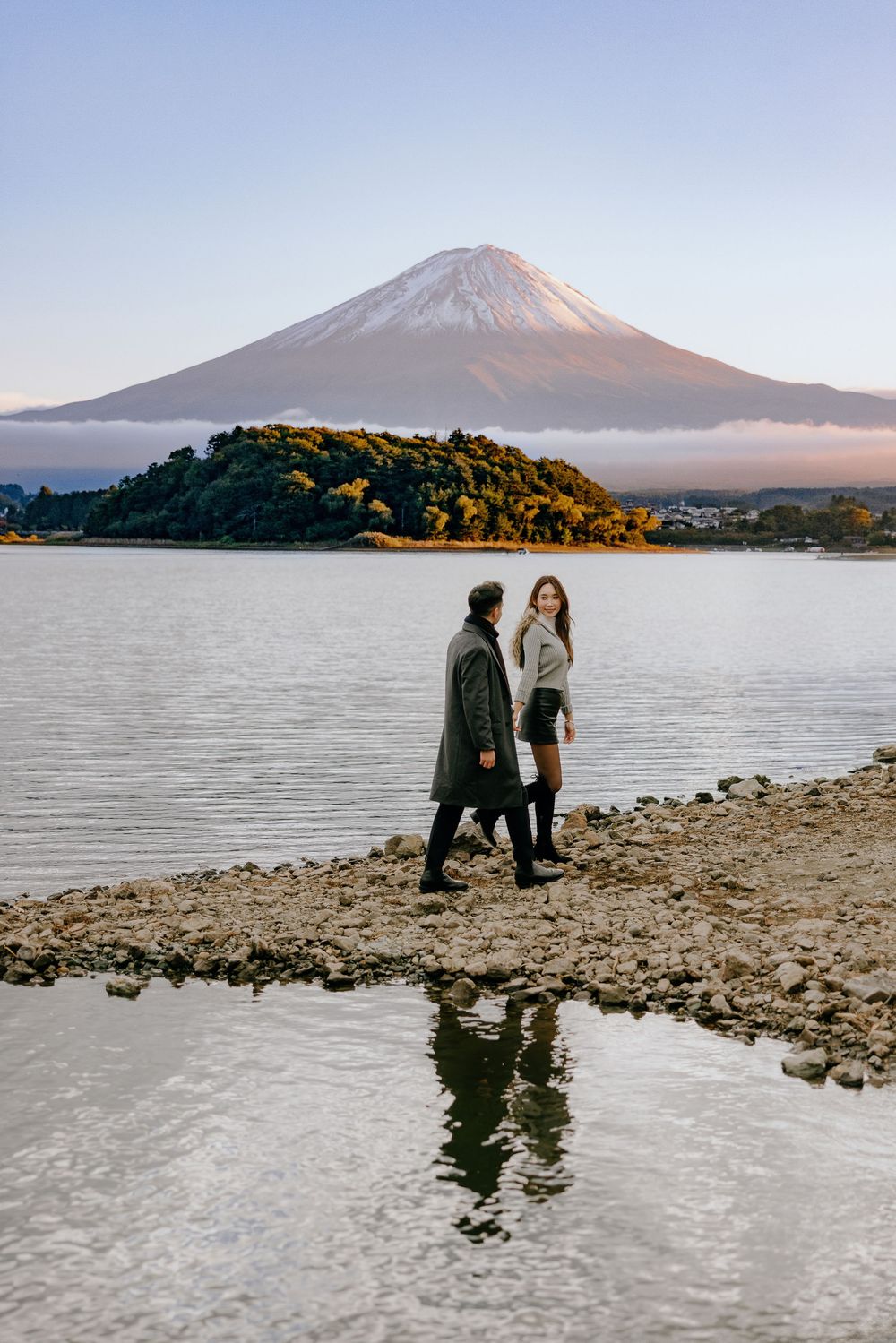 Two people stand on a rocky shoreline with Mount Fuji visible in the distance across a tranquil lake at sunset.