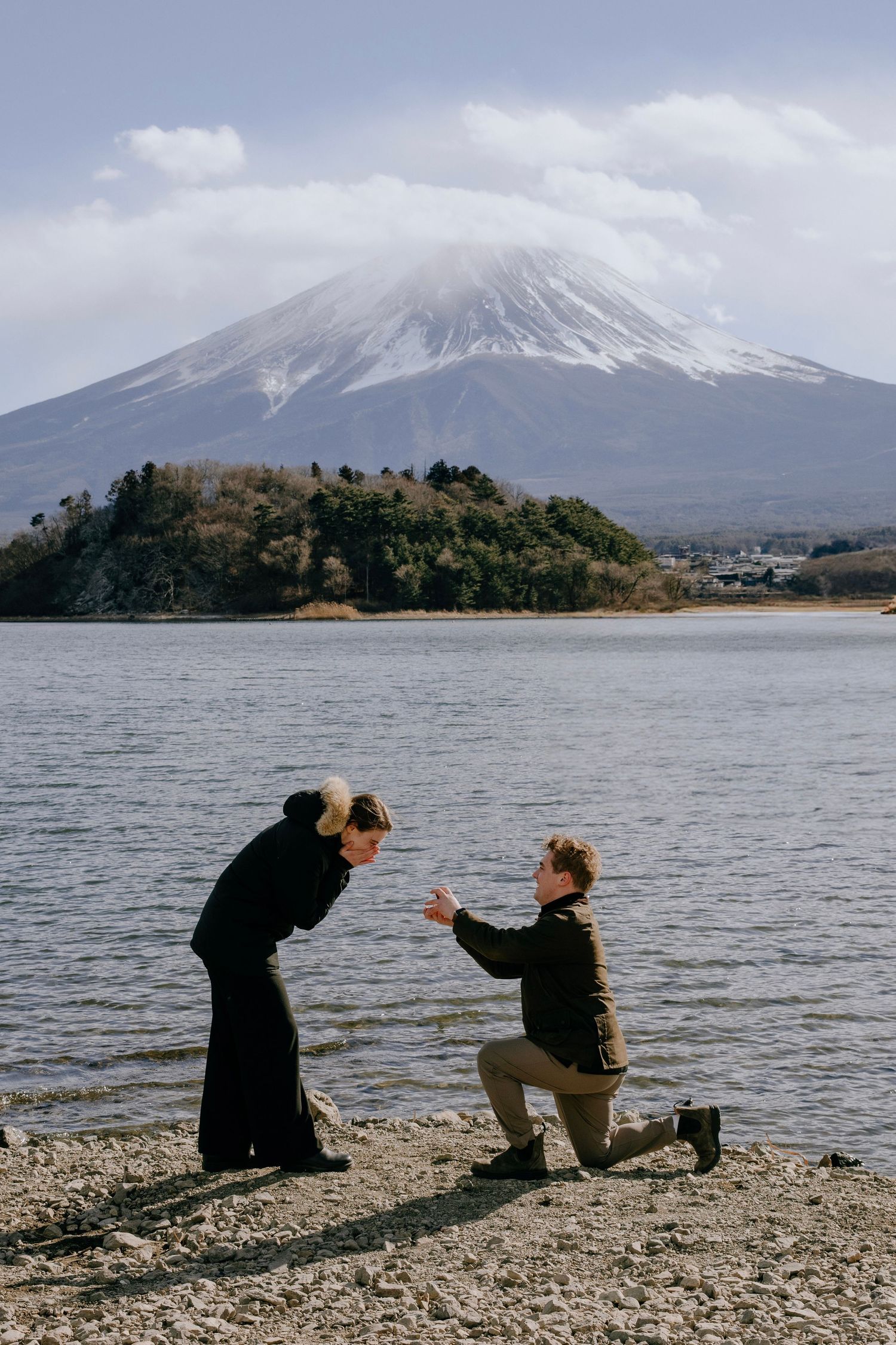 Two people sit by Lake Kawaguchiko with Mount Fuji's snow-capped peak rising majestically in the background.