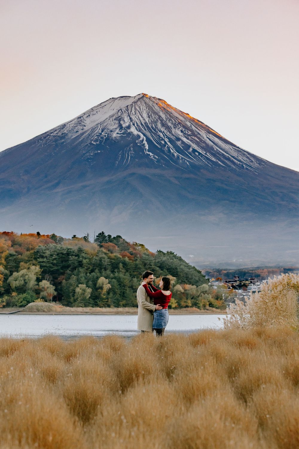 A couple embraces in a field of golden grass with Mount Fuji's snow-capped peak rising majestically in the background.
