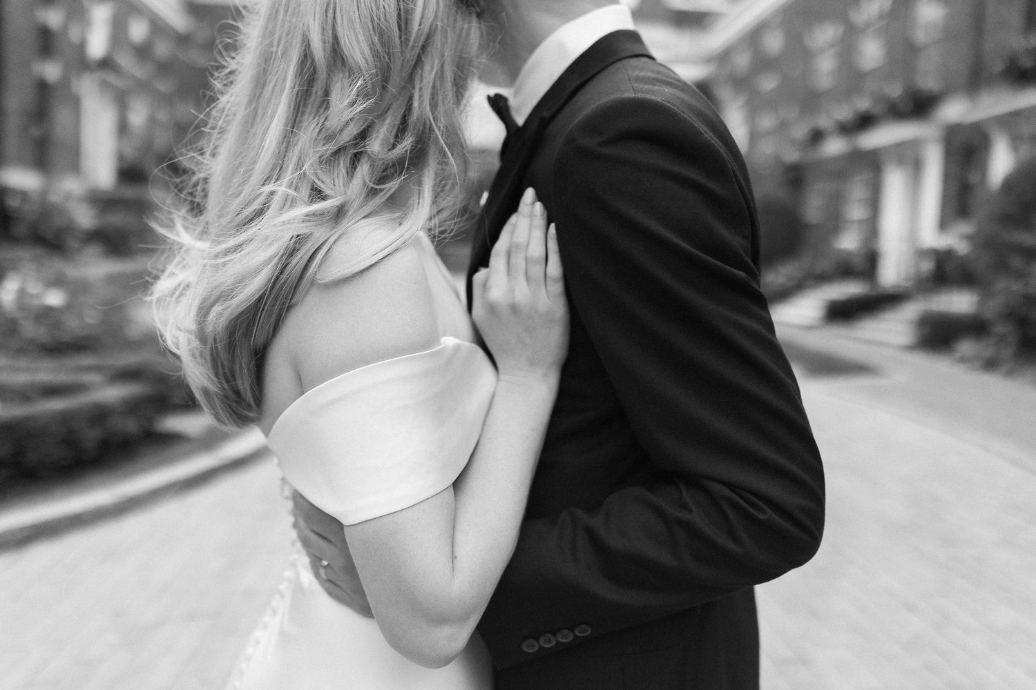 Black and white photo of a bride in off-shoulder gown embracing a groom in a tuxedo outdoors.
