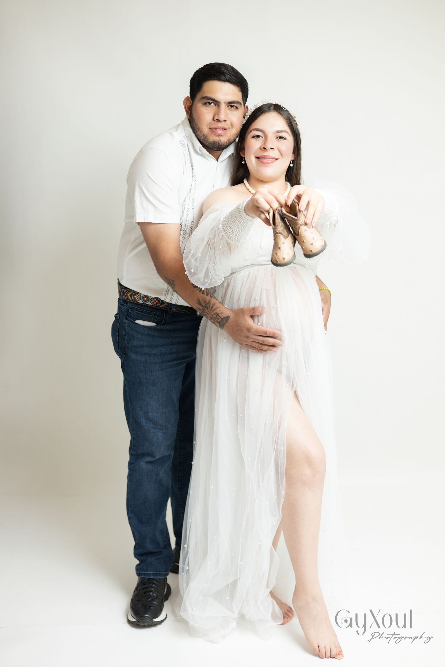 A couple in white outfits poses together against a light background in a romantic maternity photoshoot series.