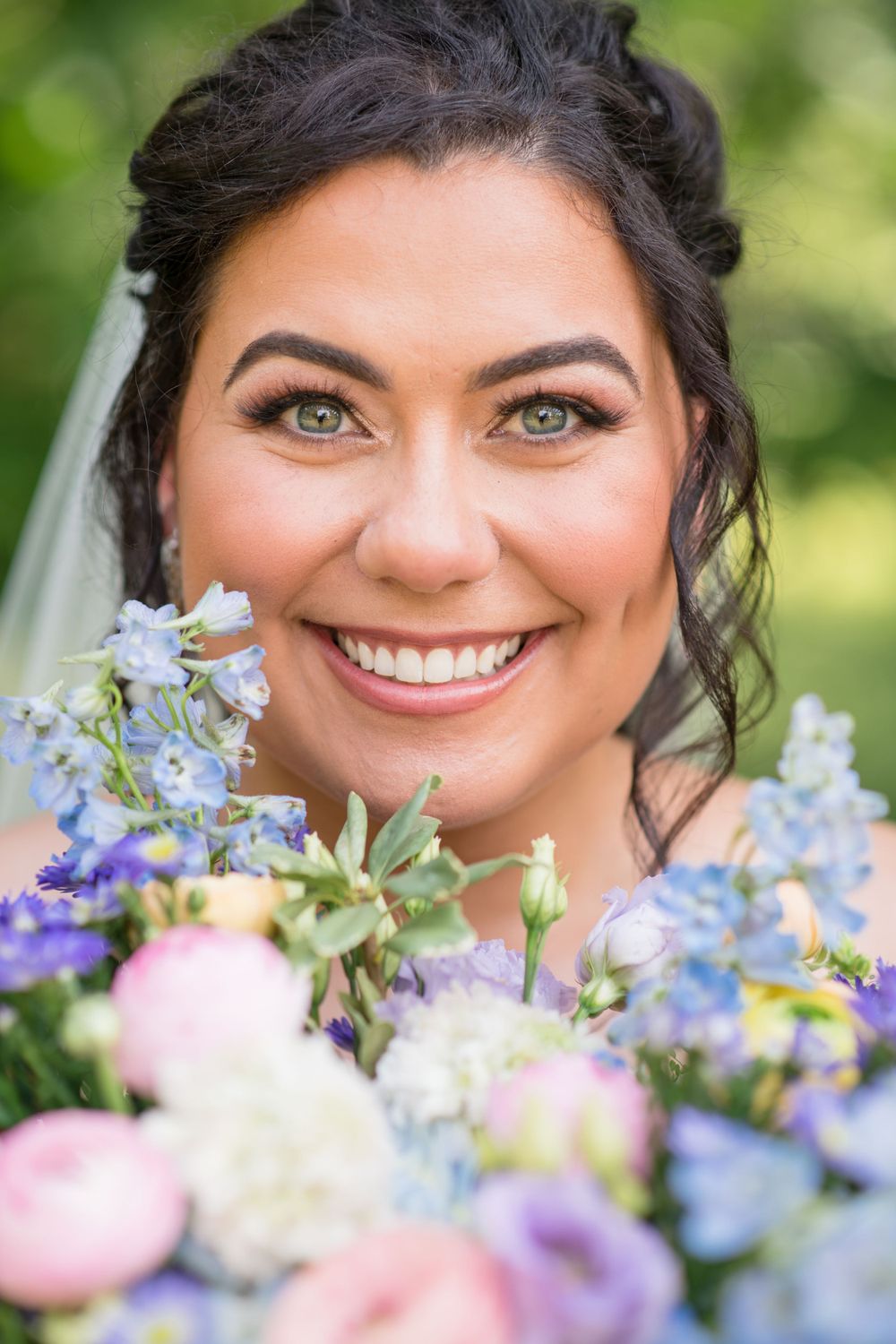 Beautiful bridal portrait closeup with delicate blue and pink bouquet flowers against a soft green outdoor background.