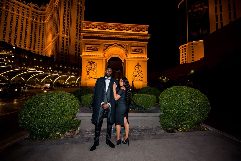 Couple standing in front of illuminated Paris Las Vegas hotel featuring replica Arc de Triomphe at night.