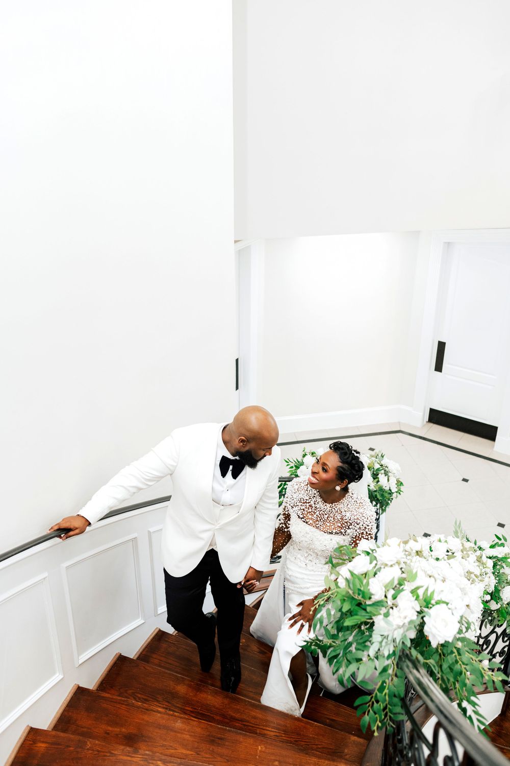 Couple in wedding attire descending white staircase with lush white floral arrangements.