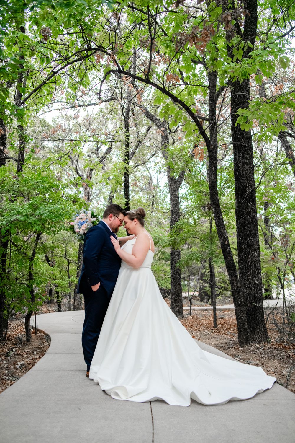 A couple shares a tender moment on a winding path surrounded by lush green trees in a forest setting.
