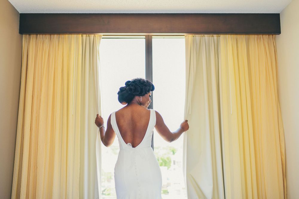A bride wearing an elegant white backless wedding dress opens yellow curtains in a bright room.