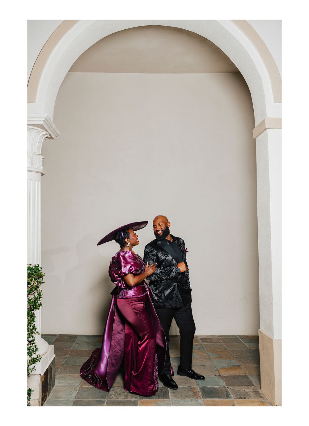 A couple poses in a white archway, wearing elegant matching outfits in burgundy and black formal attire.