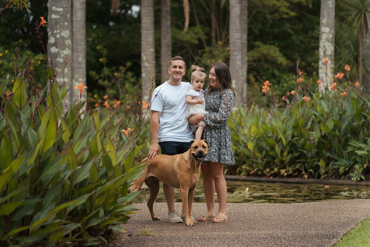 A couple and their dog with their daughter walk along a garden path surrounded by lush tropical plants and orange flowers family photoshoot